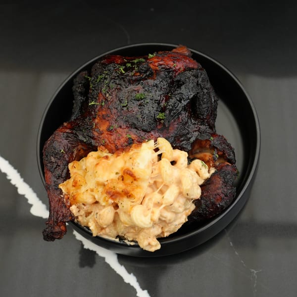 A whole roasted chicken with crispy skin and a side of creamy mac and cheese in a black bowl.
