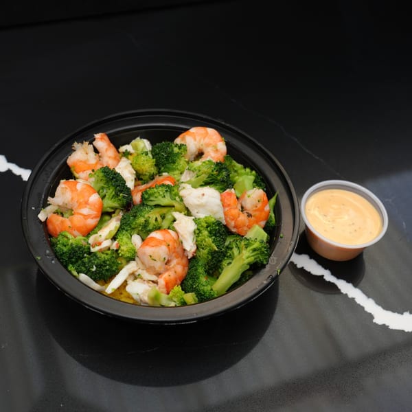Shrimp and broccoli stir-fry in a black bowl with a side of orange sauce on a dark counter.